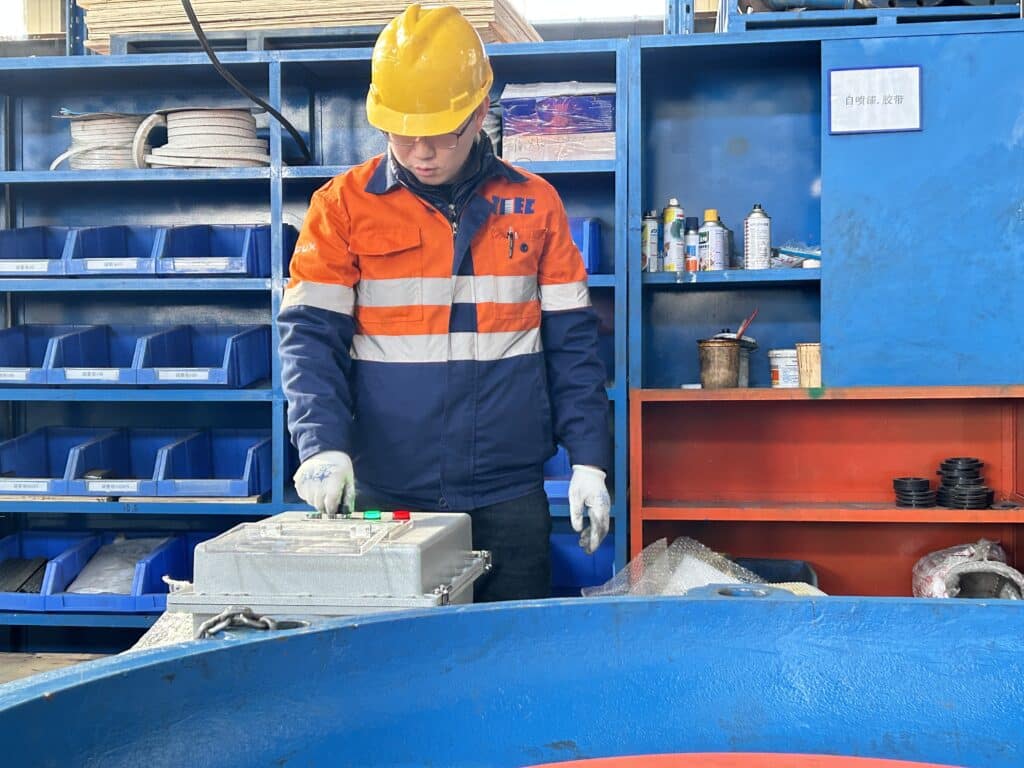 A worker wearing a TIIEC-branded uniform and a yellow hard hat operates a control panel in an industrial workspace.