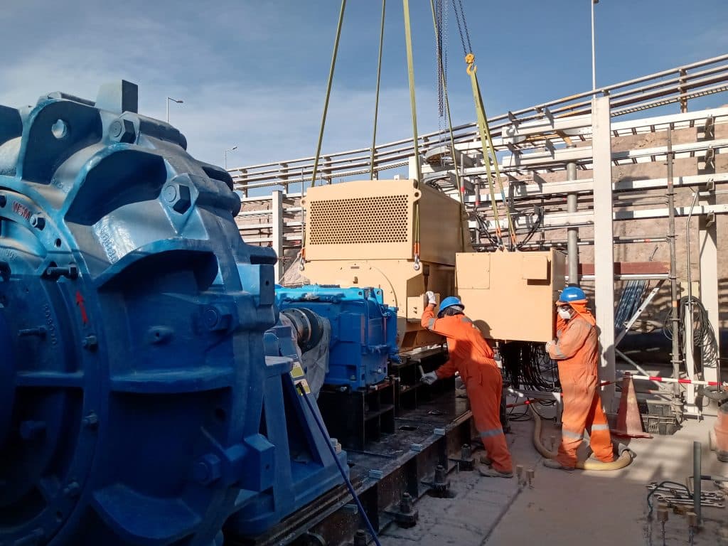 Two workers in orange safety suits and blue helmets install heavy industrial pump equipment at a facility under clear skies.